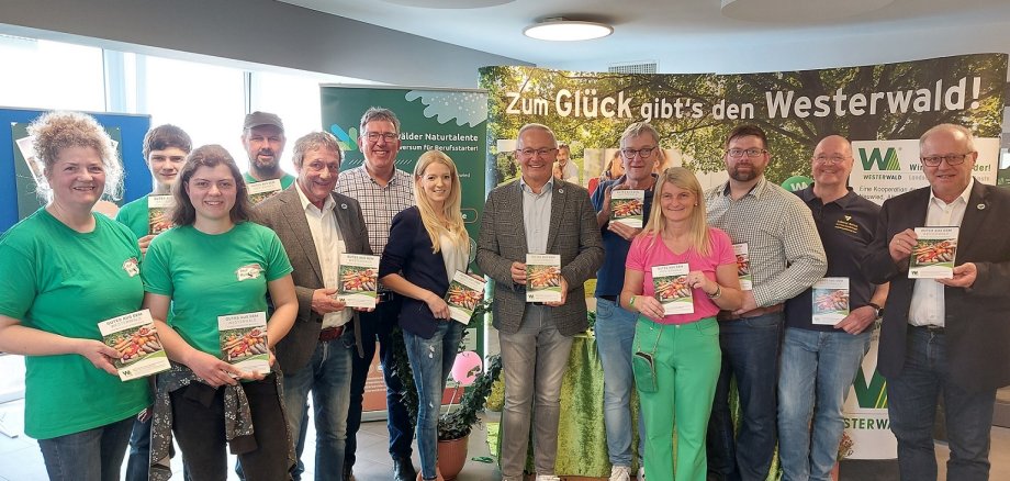 Frauen und Männer stehen mit einer Broschüre in der Hand vor einer Plakatwand.