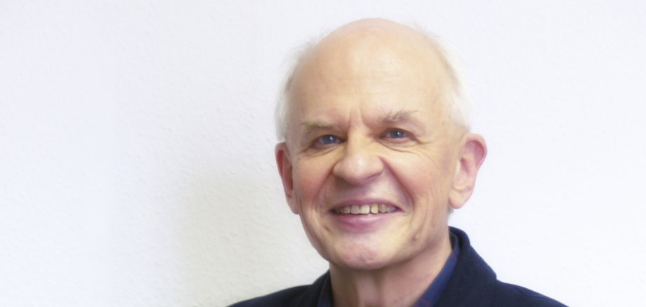 Ein älterer Herr lächelt in die Kamera und hält seine Brille in der Hand.