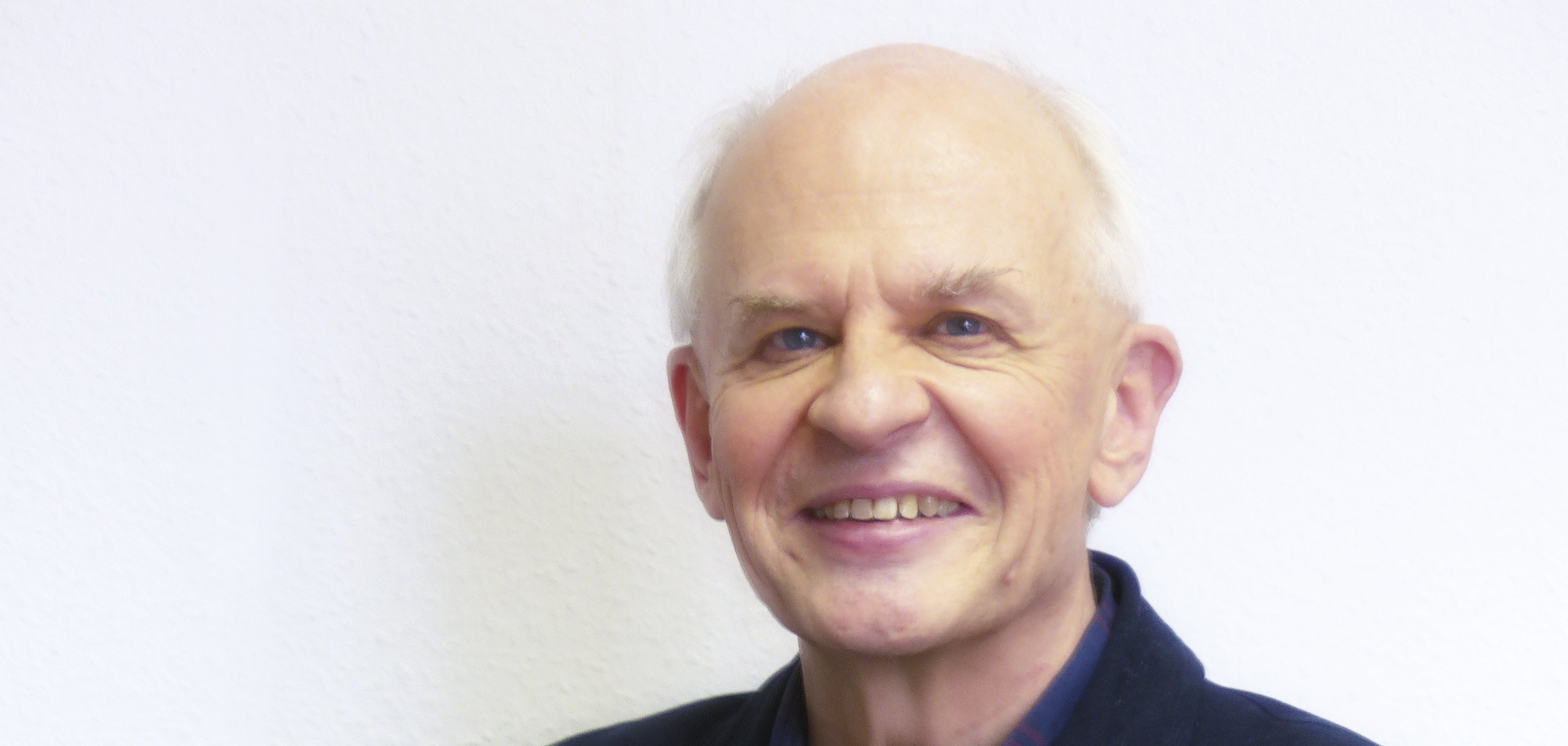 Ein älterer Herr lächelt in die Kamera und hält seine Brille in der Hand.
