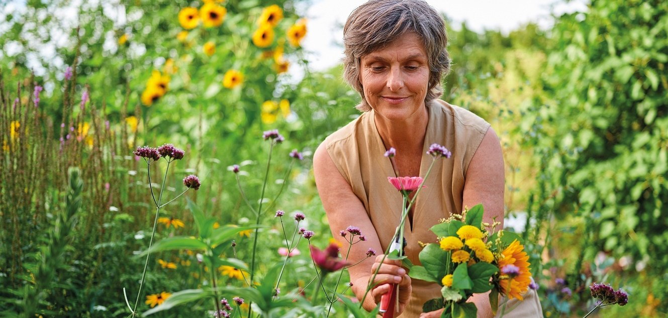 Eine Frau steht in Mitten von bunten Wildblumen und hält einen abgeschnittenen Strauß in Richtung Kamera.