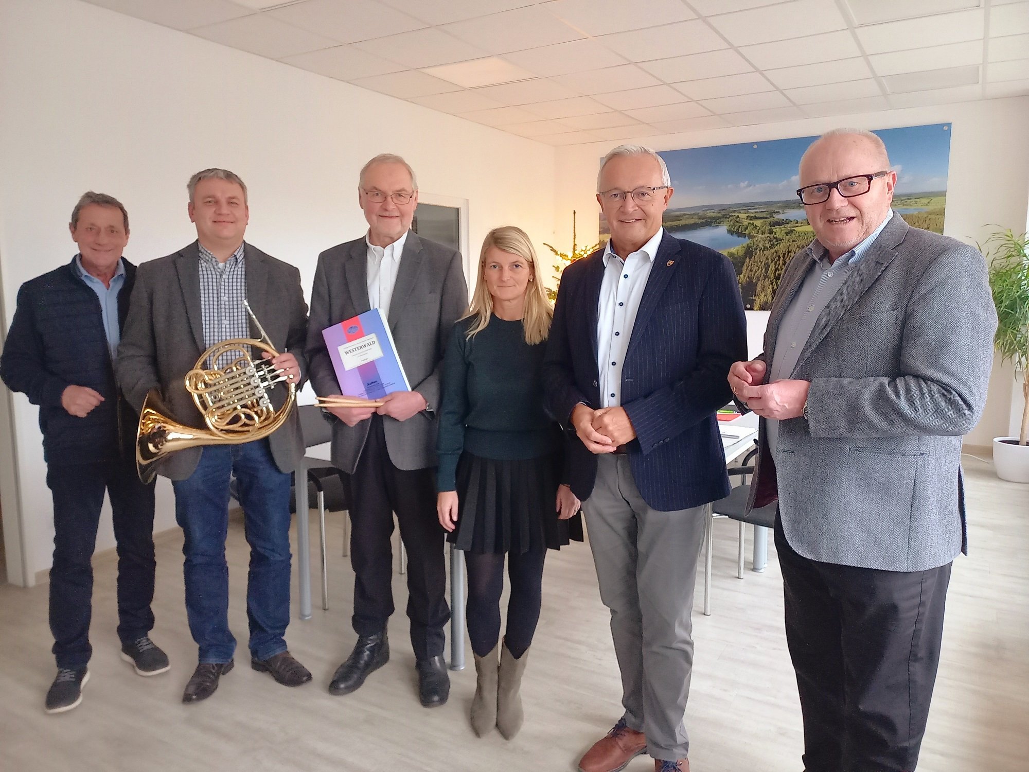 In einem Raum stehen eine Frau und fünf Männer nebeneinander, von denen einer ein Musikinstrument und ein anderer ein Notenheft in den Händen hält.