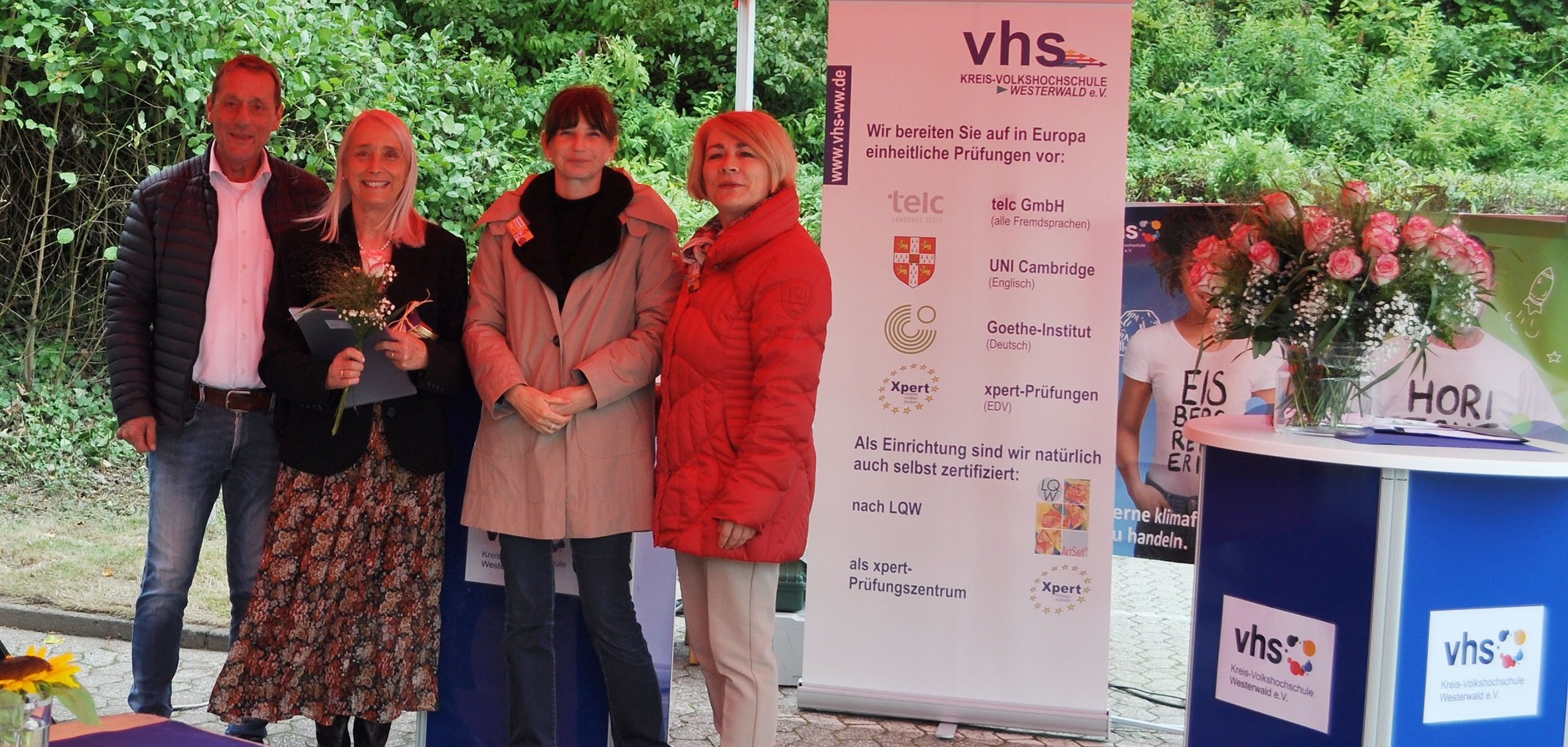 In einem Pavillon stehen neben dem Roll-up der Kreis-Volkshochschule eine Frau mit Blumen und Urkunde in der Hand, zwei weitere Frauen und der Landrat.