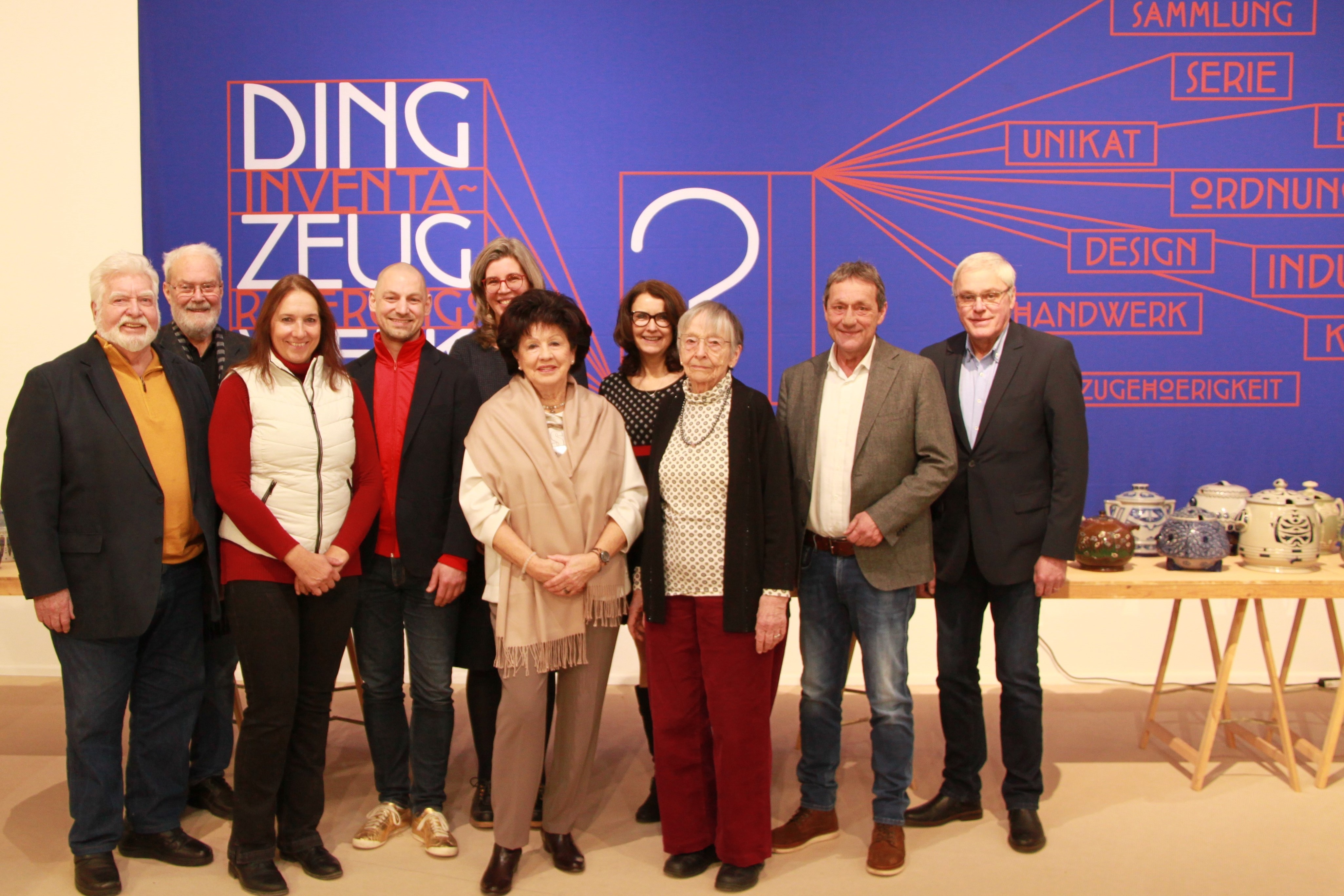 Männer und Frauen stehen vor einer blauen Plakatwand mit der Aufschrift "Ding-Zeug-Inventarisierung"