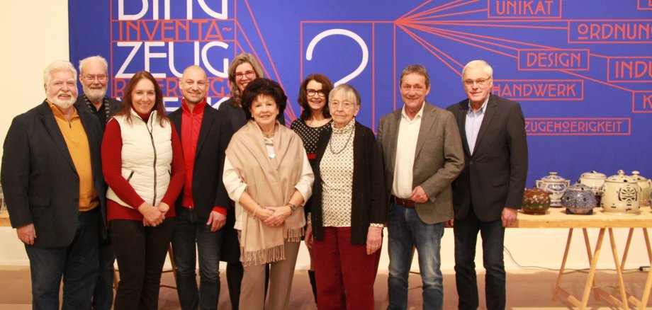Männer und Frauen stehen vor einer blauen Plakatwand mit der Aufschrift "Ding-Zeug-Inventarisierung"