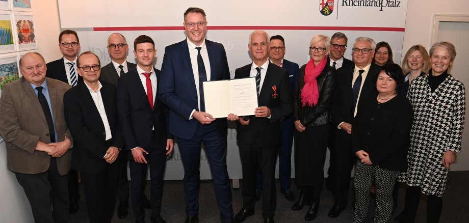 Der Sozialminister des Landes Rheinland-Pfalz überreicht das Bundesverdienstkreuz an einen Bürger aus dem Westerwaldkreis. Die beiden stehen in Mitten einer Gruppe von Menschen.