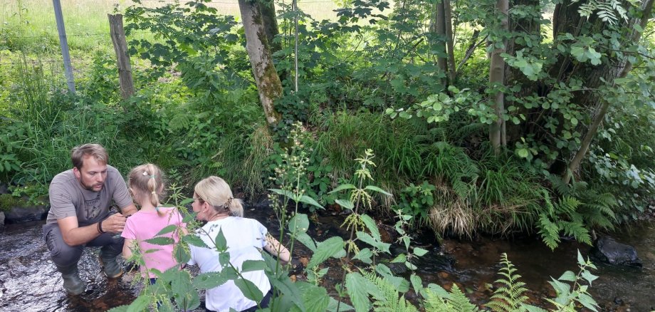 Eine Frau und ein Mann knien mit einem Mädchen an einem Bachlauf.