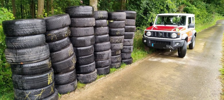 Sechs Reifenstapel mit je sieben Reifen sind mitten im Wald an einem Weg aufgestapelt. Im Hintergrund steht ein Auto der Kreisverwaltung.