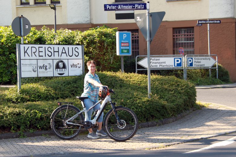 Eine Radfahrerin steht neben ihrem Fahrrad vor einem Schild der Kreisverwaltung.