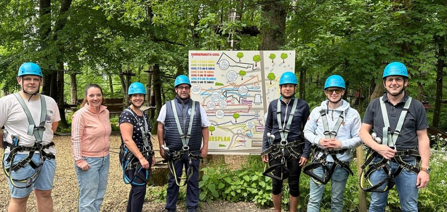 Männer und Frauen mit Helmen auf dem Kopf stehen vor einer Infotafel im Kletterwald.