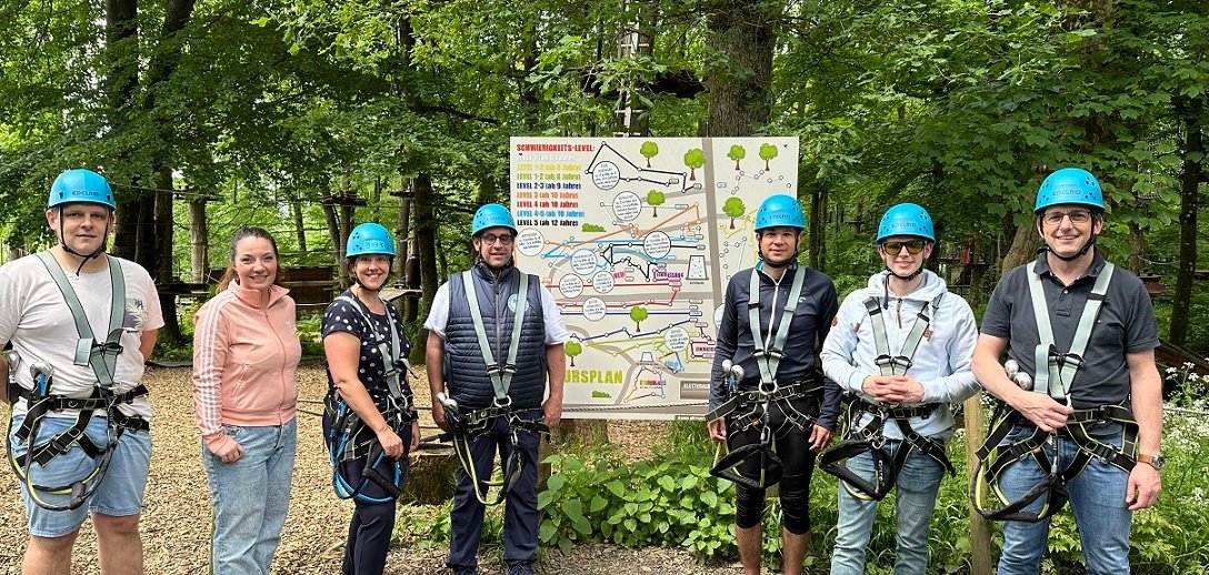 Männer und Frauen mit Helmen auf dem Kopf stehen vor einer Infotafel im Kletterwald.