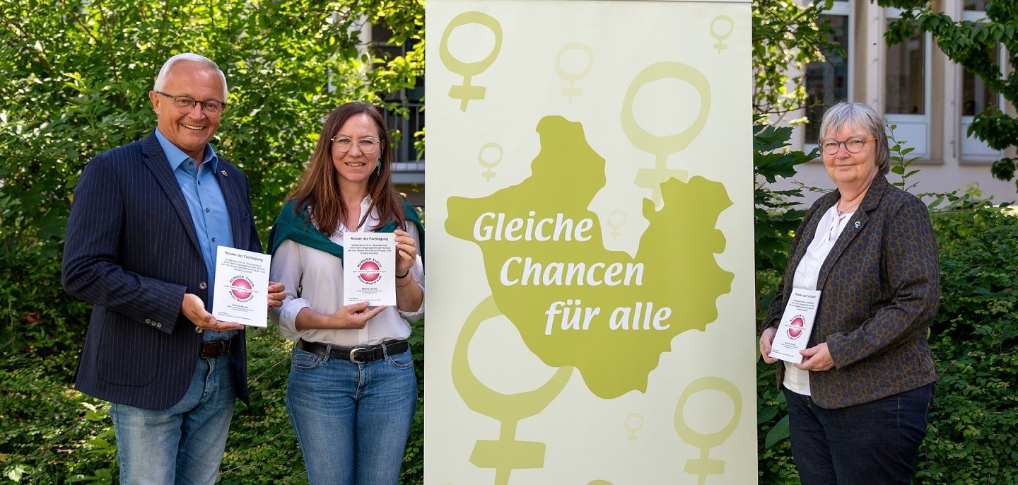Neben einem Banner mit der Aufschrift "Gleiche Chancen für alle" stehen zwei Frauen und ein Mann mit einem gedruckten Reader in der Hand.