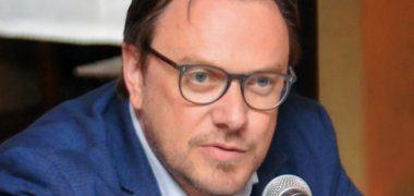 In ein Mikrofon sprechend sitzt ein Mann mit Brille und blauem Jacket an einem Tisch.