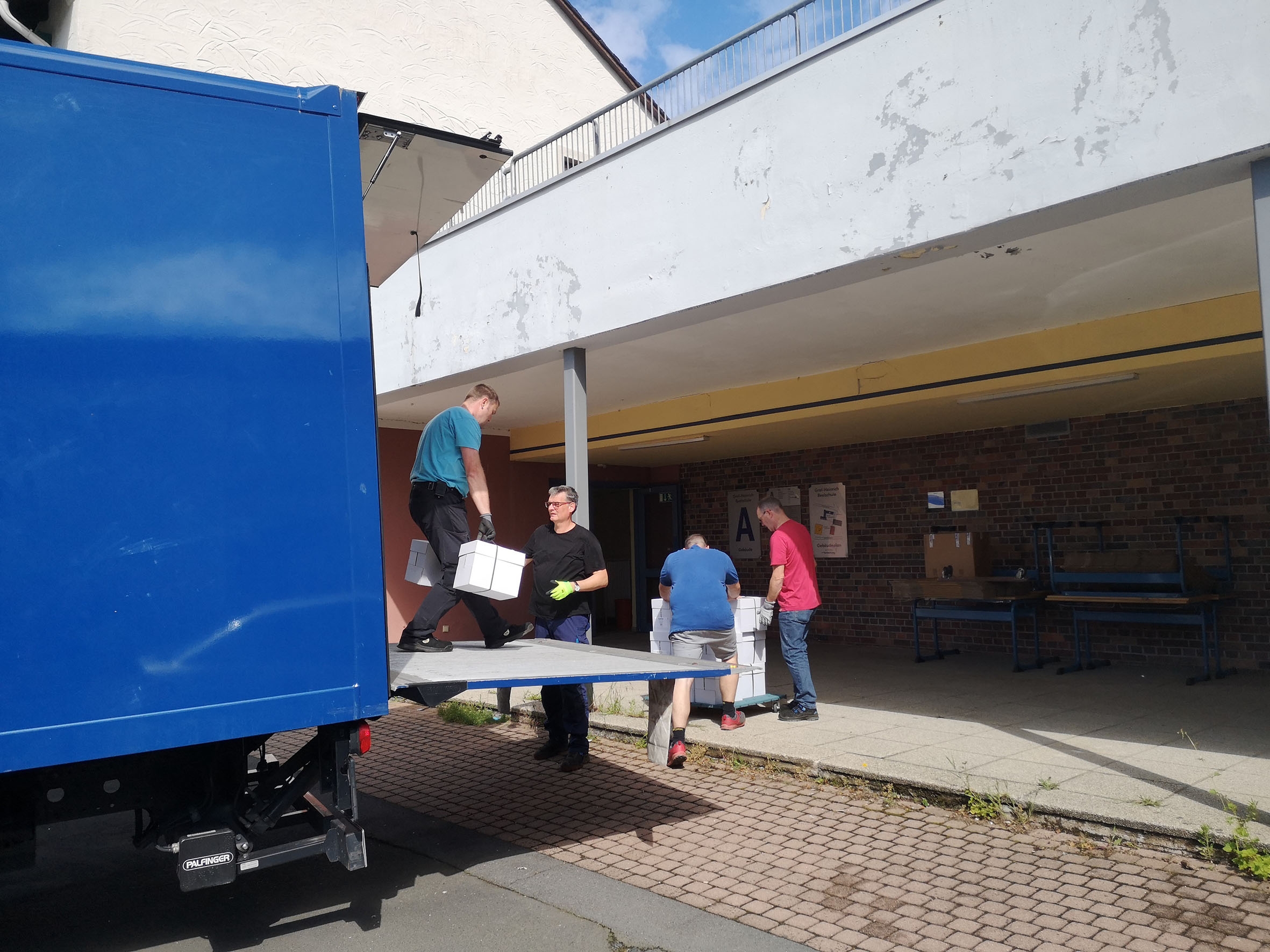 Von einem blauen Lkw laden vier Männer weiße Kartons vor einer Schule ab.