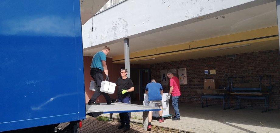 Von einem blauen Lkw laden vier Männer weiße Kartons vor einer Schule ab.