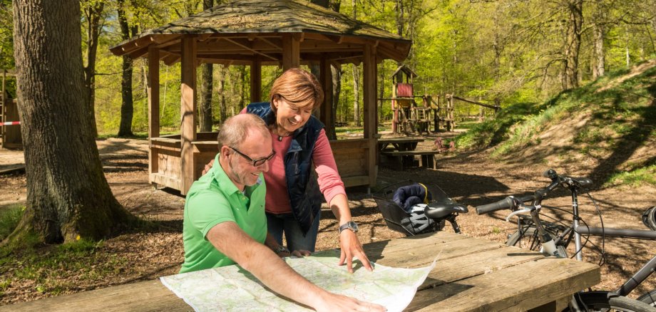Ein sitzender Mann und eine stehende Frau schauen gemeinsam auf eine Karte. Diese liegt auf einem Holztisch an einem Wanderrastplatz, an den ihre Fahrräder gelehnt sind.