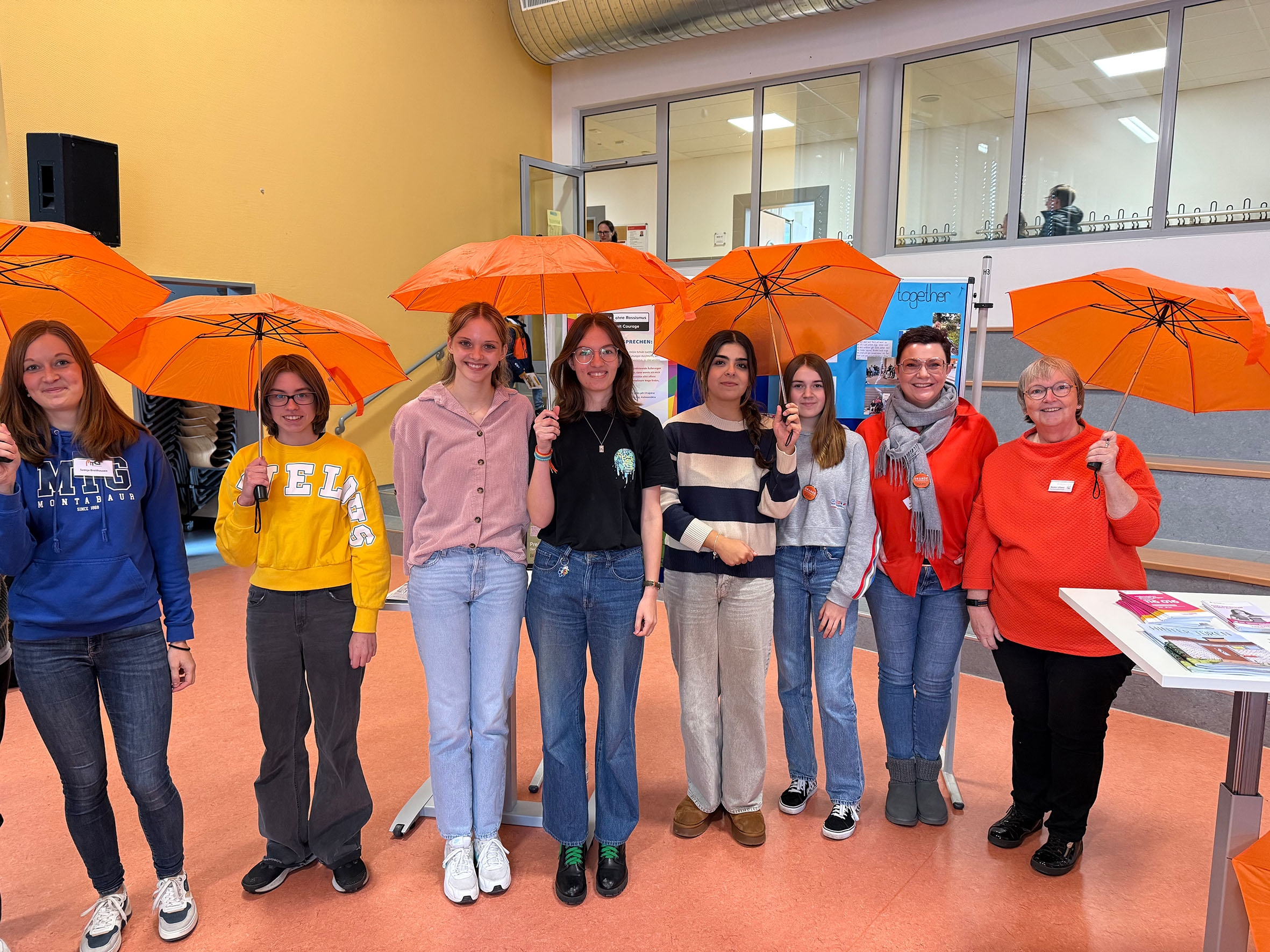 Acht Frauen lachen mit orangenen Regenschirmen in der Hand in die Kamera.