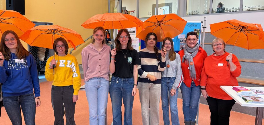 Acht Frauen lachen mit orangenen Regenschirmen in der Hand in die Kamera.