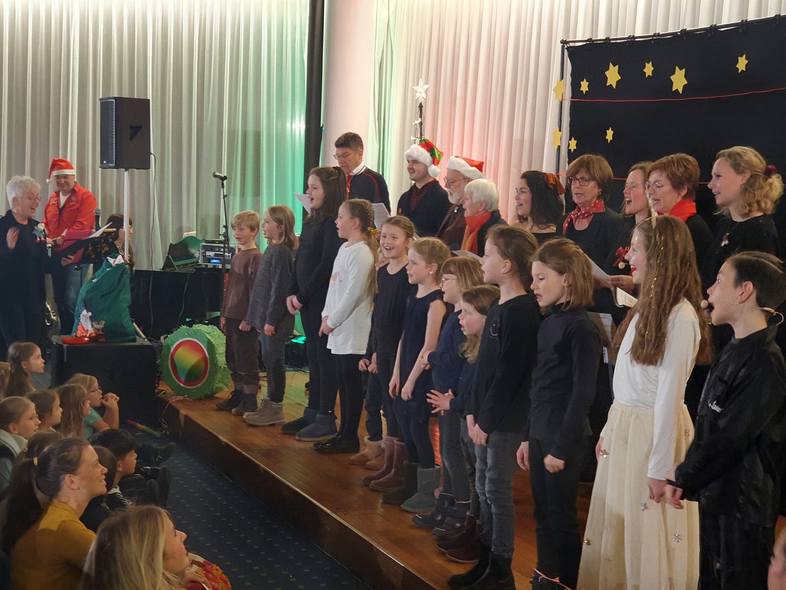 Kinder und Erwachsene singen zum Teil weihnachtlich gekleidet gemeinsam auf einer Bühne.