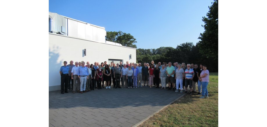 Eine große Gruppe Menschen steht im Halbkreis vor einem Gebäude und Bäumen.