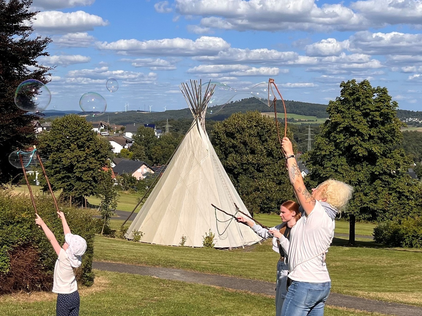 Ein Kind und zwei Frauen stehen vor einem Tipi auf einer Wiese und machen riesige Seifenblasen.