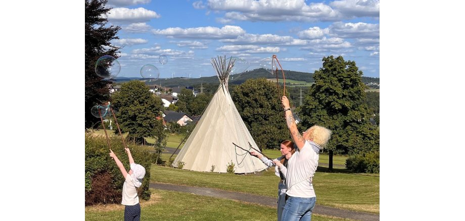 Ein Kind und zwei Frauen stehen vor einem Tipi auf einer Wiese und machen riesige Seifenblasen.