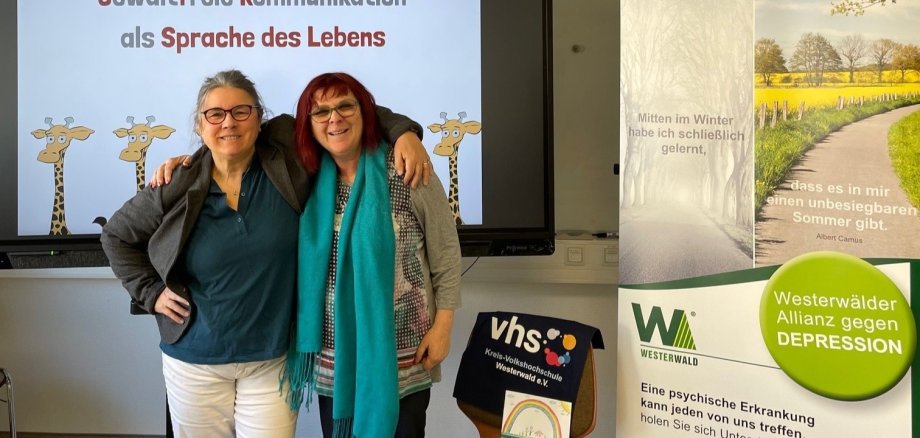 Eine Frau hat einer anderen den Arm um die Schultern gelegt, beide stehen vor einer Leinwand.