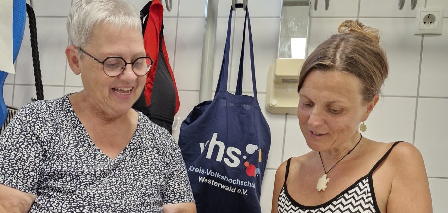 Zwei Frauen sitzen in einem Raum beieinander und die ältere erklärt der jüngeren etwas im Programmheft der vhs.