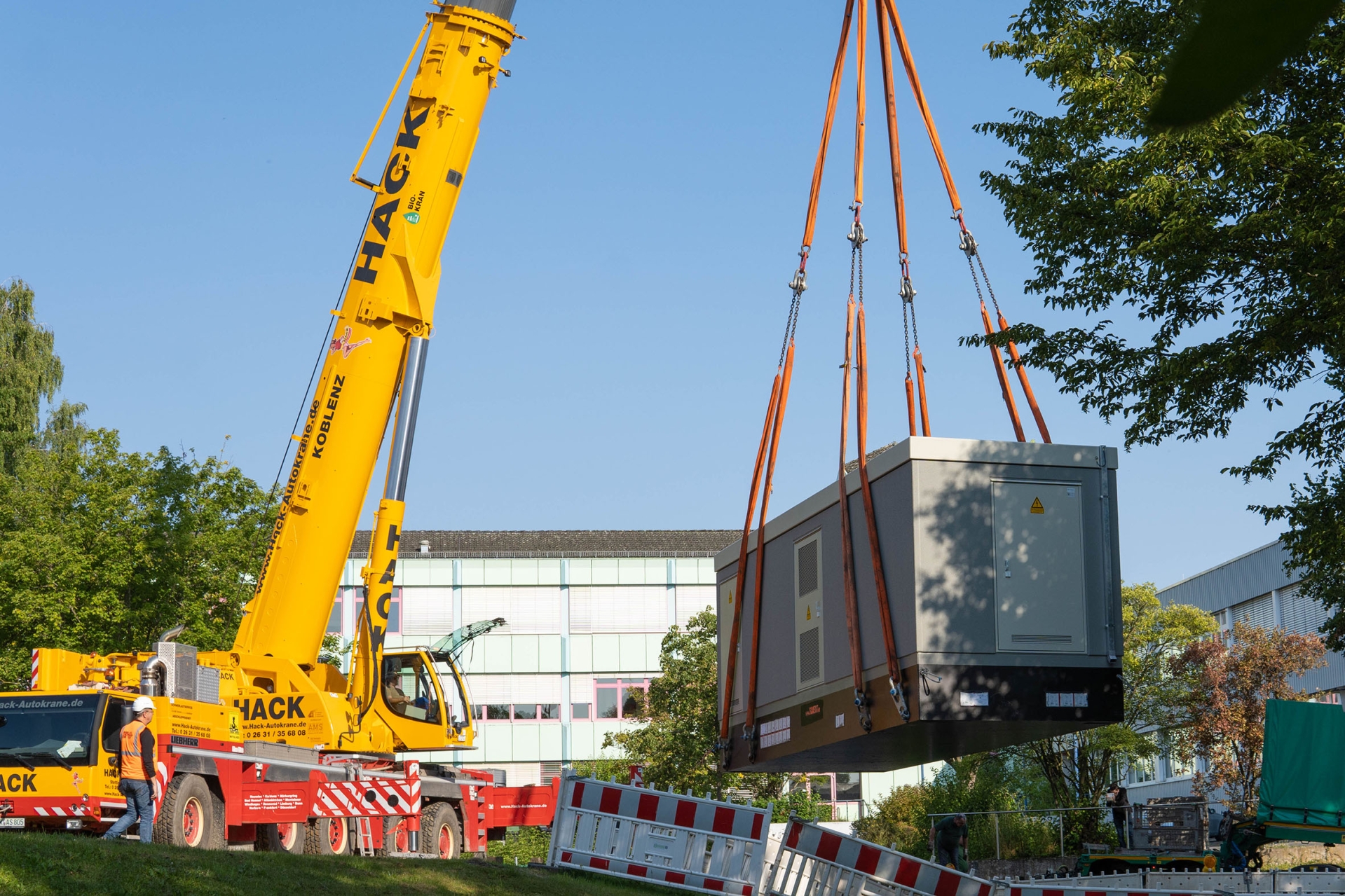 An einem gelben Kran vor einem Schulgebäude hängt eine Trafostation.