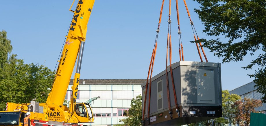 An einem gelben Kran vor einem Schulgebäude hängt eine Trafostation.