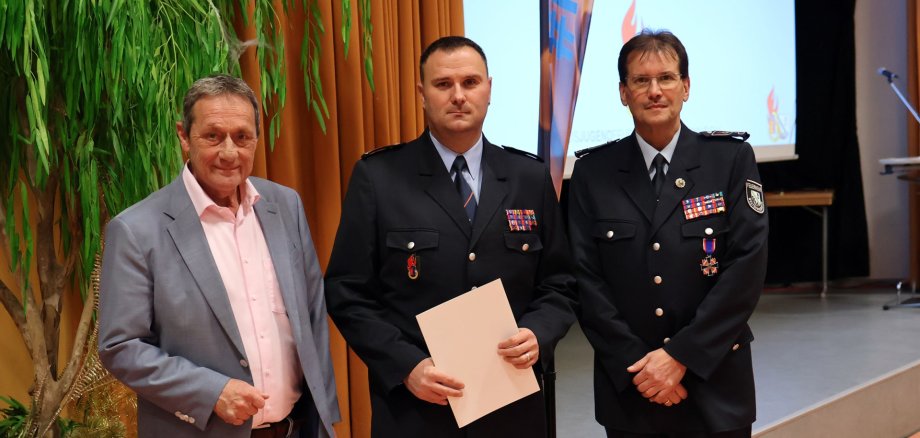 Drei Männer, der Landrat im grauen Anzug und Bach sowie Krekel in Dienstuniform stehen vor einer Bühne.