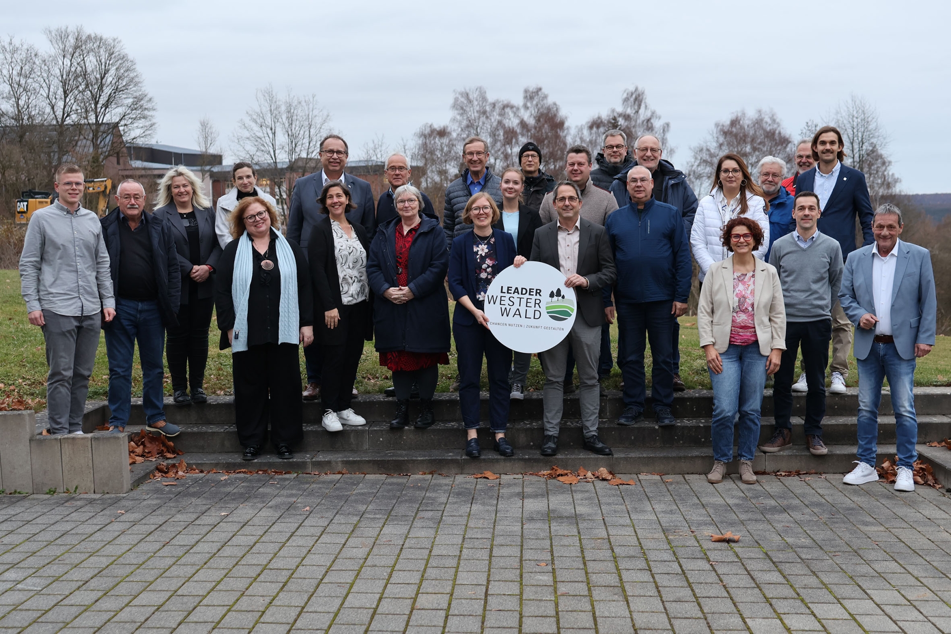 Mehrere Frauen und Männer stehen auf drei Stufen vor einer Wiese, in der Mitte halten zwei Personen ein Schild mit der Aufschrift Leader Westerwald.
