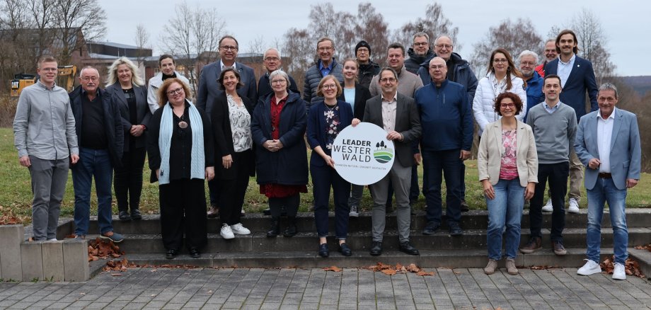Mehrere Frauen und Männer stehen auf drei Stufen vor einer Wiese, in der Mitte halten zwei Personen ein Schild mit der Aufschrift Leader Westerwald.
