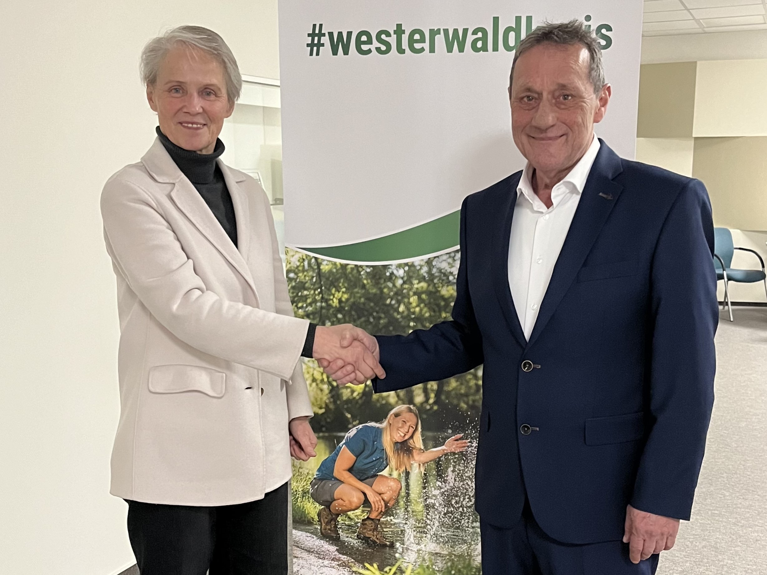 Eine Frau im Blazer gratuliert dem Landrat per Handschlag vor einem Rollup des Westerwaldkreises.