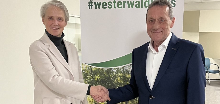 Eine Frau im Blazer gratuliert dem Landrat per Handschlag vor einem Rollup des Westerwaldkreises.