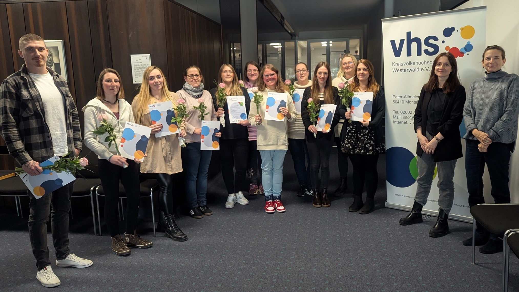 Vor einem Roll-up der Kreis-Volkshochschule stehen mit Blumen und Urkunden in den Händen die Teilnehmenden der Fortbildung.