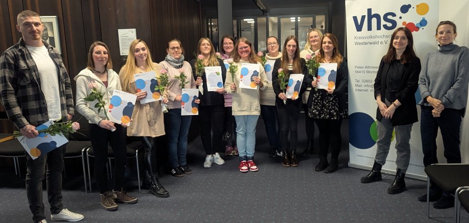 Vor einem Roll-up der Kreis-Volkshochschule stehen mit Blumen und Urkunden in den Händen die Teilnehmenden der Fortbildung.