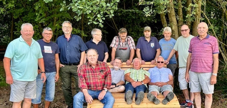 Mehrere Männer sitzen auf einer großen Holzbank oder stehen daneben.