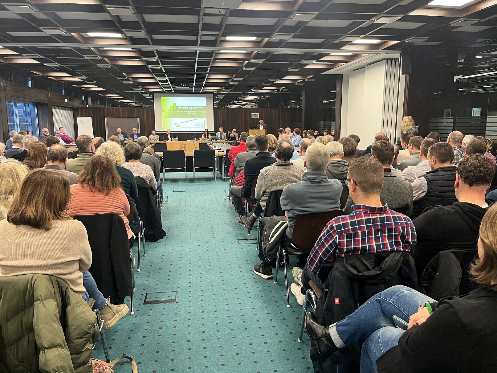 In einem großen Versammlungssaal sitzen viele Menschen in Stuhlreihen. Vorn steht der Landrat am Rednerpult vor einer Powerpointpräsentation.
