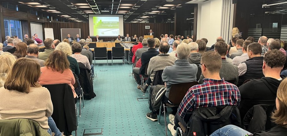 In einem großen Versammlungssaal sitzen viele Menschen in Stuhlreihen. Vorn steht der Landrat am Rednerpult vor einer Powerpointpräsentation.