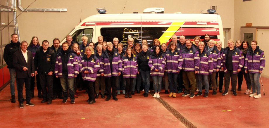 Eine große Gruppe Menschen steht in lila Jacken vor einem Rettungswagen.