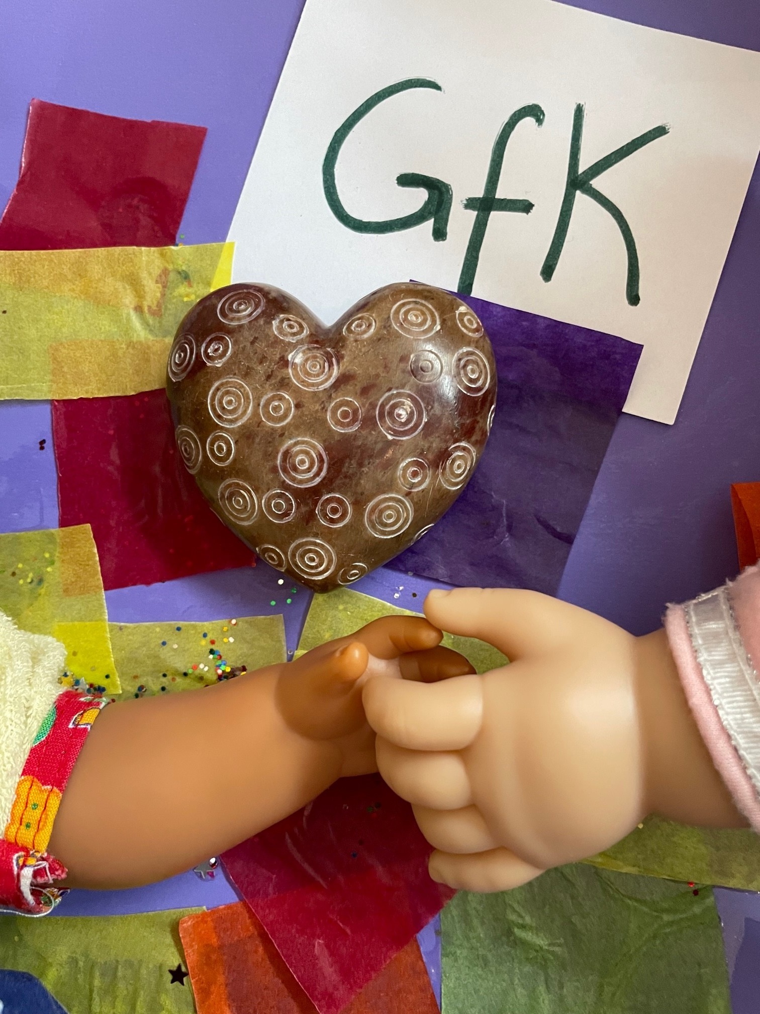 Auf buntem Papier mit der Aufschrift GfK liegen ein Keramikherz und zwei Puppen, deren Hände sich berühren.