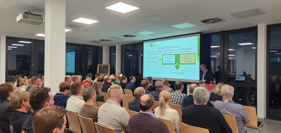 Ein Veranstaltungsraum mit voll besetzten Stuhlreihen und einem Vortragenden vorn.