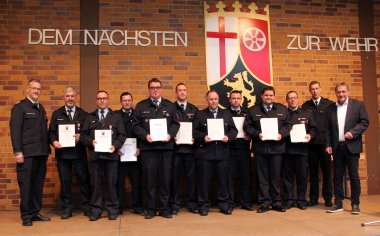 Neun Männer in blauer Feuerwehr-Dienstuniform mit einer weißen Urkunde in der Hand stehen eingerahmt von Landrat und den beiden BKIs auf einer Bühne.
