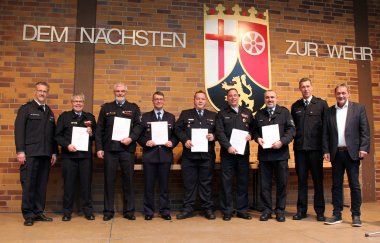Sechs Männer in blauer Feuerwehr-Dienstuniform mit einer weißen Urkunde in der Hand stehen eingerahmt von Landrat und den beiden BKIs auf einer Bühne.