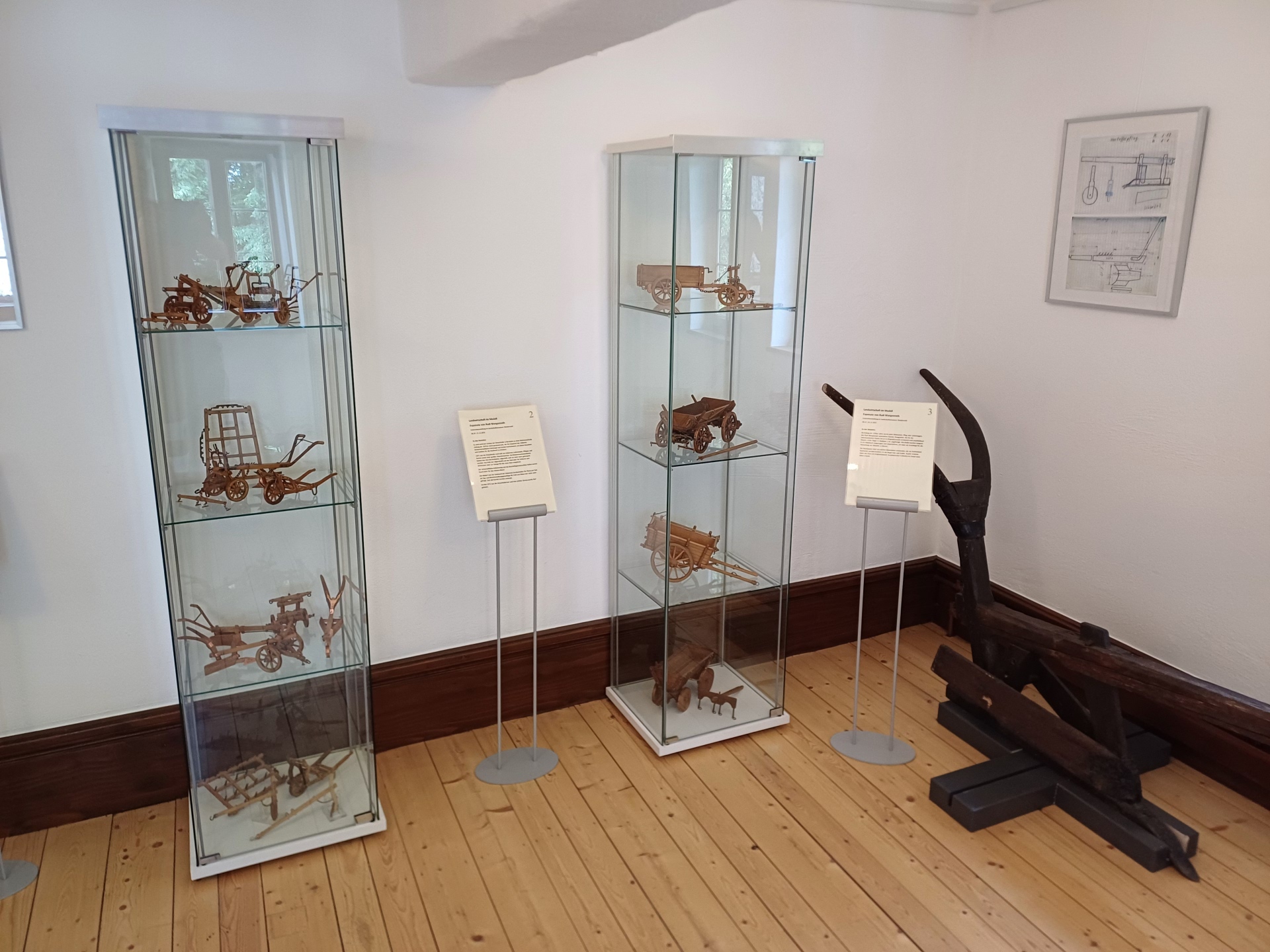 Verschiedenste Miniatur-Modelle von landwirtschaftlichen Geräten wie ein Pflug oder ein Karren stehen in zwei Glasvitrinen in einem Museumsraum.