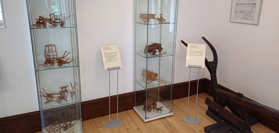 Verschiedenste Miniatur-Modelle von landwirtschaftlichen Geräten wie ein Pflug oder ein Karren stehen in zwei Glasvitrinen in einem Museumsraum.