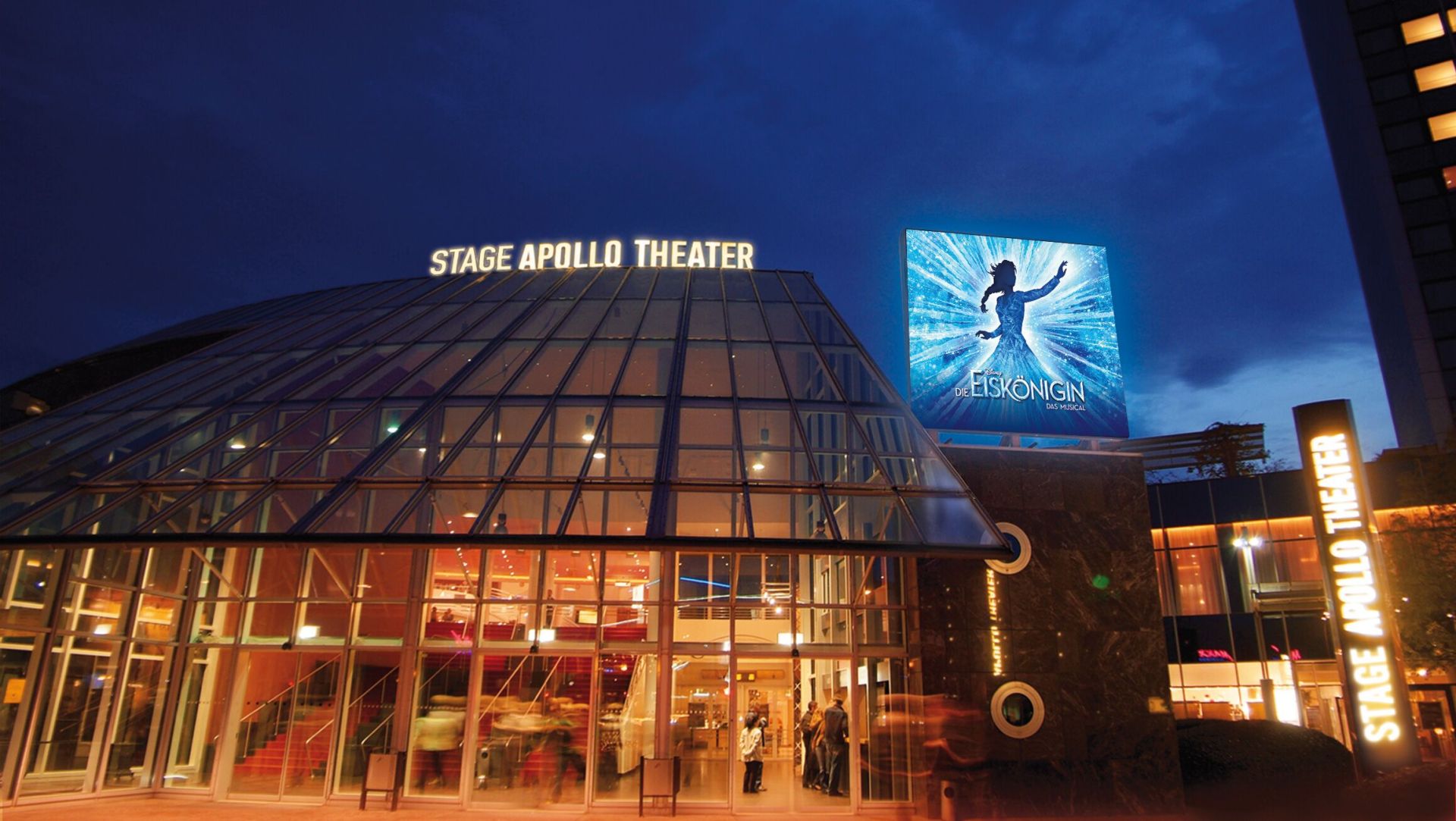 Das Stage Apollo Theater bei Nacht ist ein hell erleuchteter Glasbau, rechts mit dem Werbeplakat für das Musical "Die Eiskönigin".