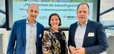 Zwei Männer und eine Frau stehen vor dem Vortragsmonitor und lächeln in die Kamera