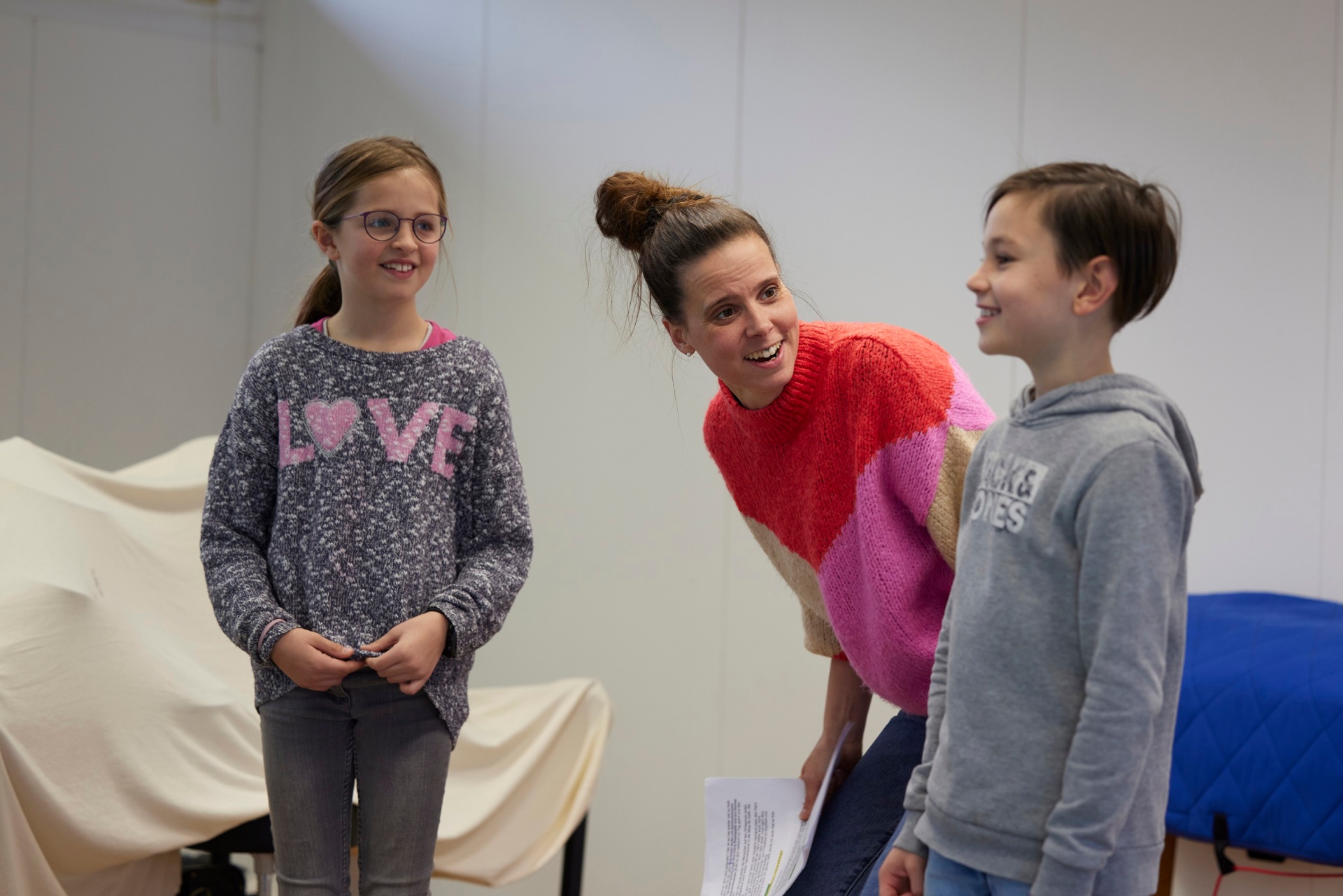 Eine Lehrerin probt mit einem lächelnden Jungen und einem Mädchen in einem Raum ein Stück ein.