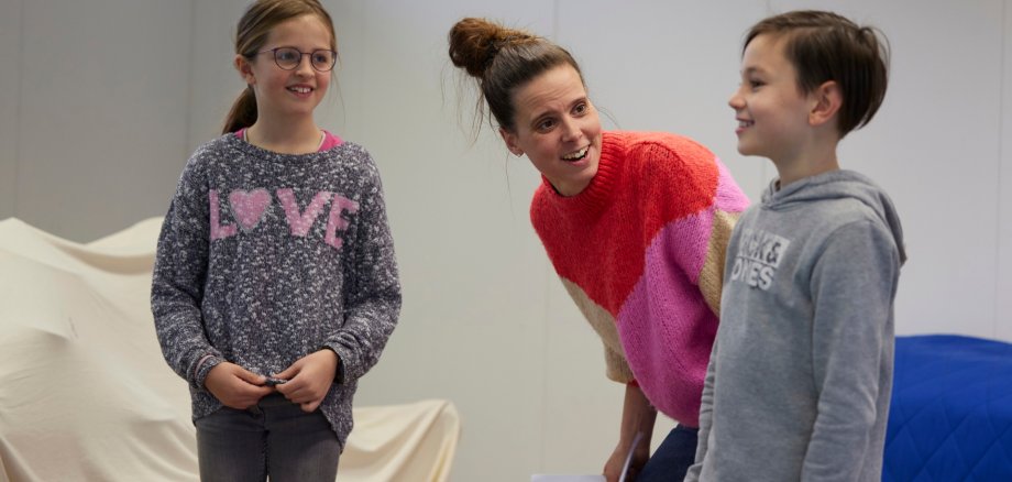 Eine Lehrerin probt mit einem lächelnden Jungen und einem Mädchen in einem Raum ein Stück ein.